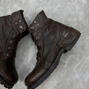 May include: A pair of brown leather boots with laces and metal eyelets. The boots have a worn, distressed appearance, with visible creases and scuffs. The soles are black and rugged, suggesting durability and a vintage style.