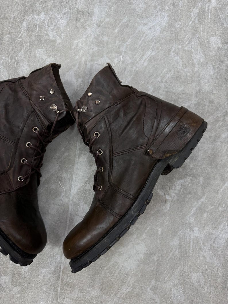 May include: A pair of brown leather boots with laces and metal eyelets. The boots have a worn, distressed appearance, with visible creases and scuffs. The soles are black and rugged, suggesting durability and a vintage style.
