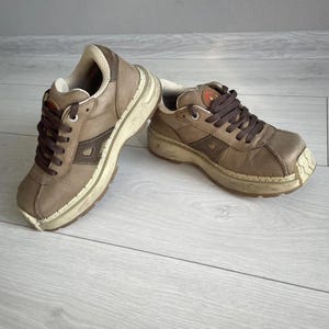 Puede incluir: Un par de zapatillas de cuero marrón con suelas blancas y cordones marrones. Las zapatillas tienen un aspecto desgastado y están hechas de cuero texturizado.