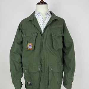 Könnte beinhalten: Olivgrüne Jacke mit mehreren Vordertaschen und Knopfverschlüssen. Ein farbenfroher Aufnäher befindet sich auf dem linken Ärmel. Die Jacke wird auf einer Schaufensterpuppe mit einem weißen Hemd mit Muster darunter präsentiert.