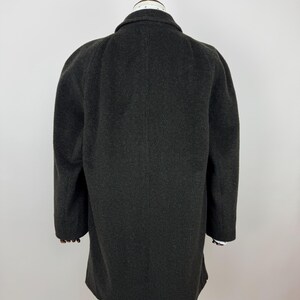 May include: A dark gray wool coat with a classic collar and long sleeves. The coat is displayed on a mannequin, showcasing its tailored fit and textured fabric. The sleeves reveal a glimpse of a white and black striped cuff.