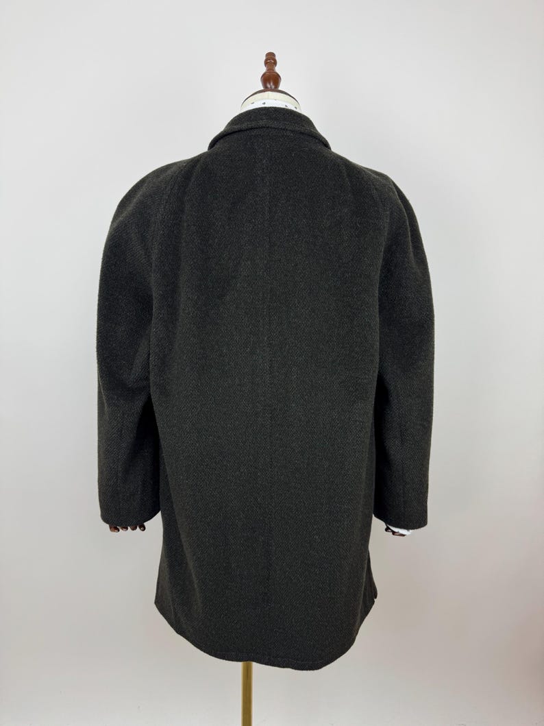 May include: A dark gray wool coat with a classic collar and long sleeves. The coat is displayed on a mannequin, showcasing its tailored fit and textured fabric. The sleeves reveal a glimpse of a white and black striped cuff.