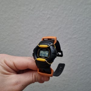 May include: Black and yellow digital wristwatch with a black band. The watch face displays the time, date, and day of the week. The watch is water resistant to 100 meters.