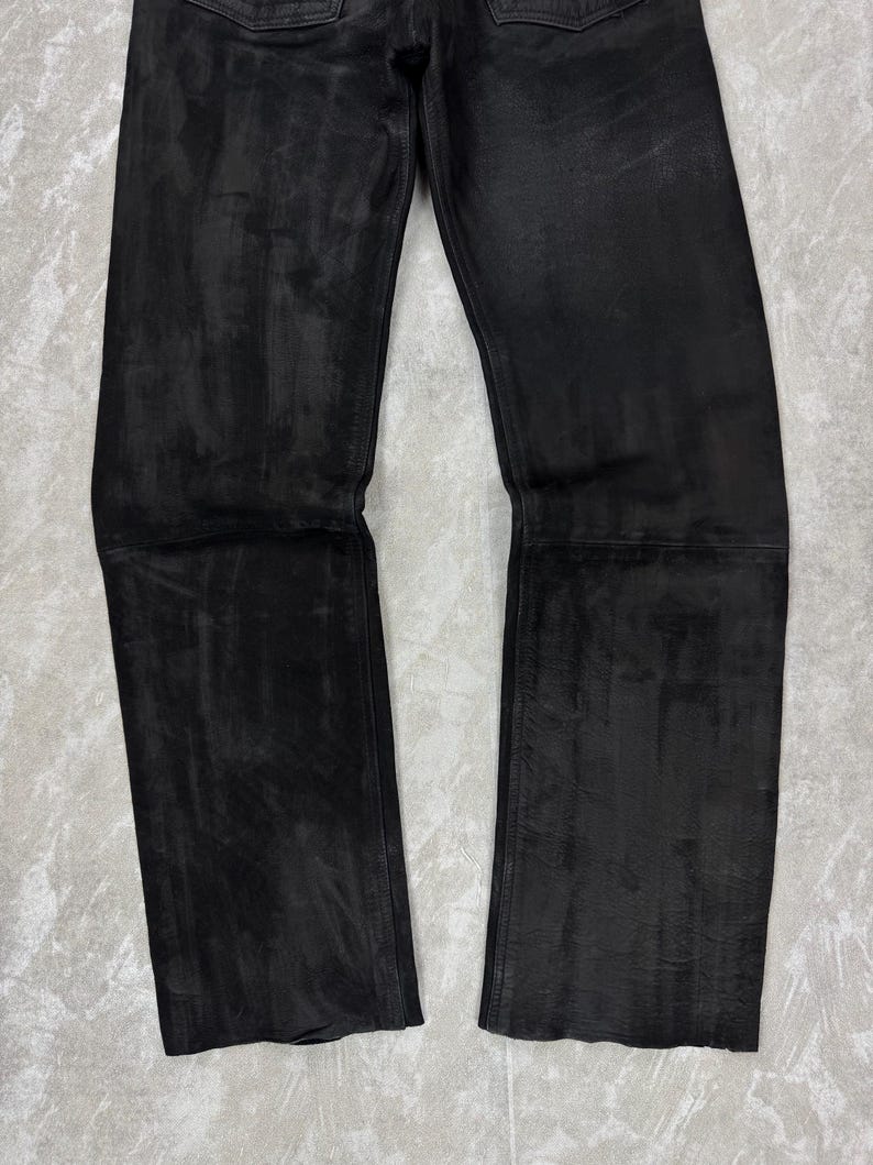 May include: A pair of black leather trousers featuring a matte finish with visible tonal variations and horizontal seam detailing across the mid-thigh area. The trousers have a straight-leg cut with a clean hem and a structured, multi-panel construction.