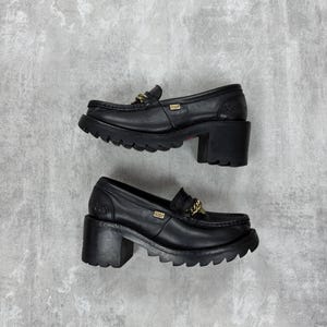 Puede incluir: Par de mocasines negros de plataforma de cuero con tacones gruesos. Cada zapato presenta un detalle de cadena dorada en el empeine y una suela con dibujo. Los zapatos se muestran sobre un fondo gris.