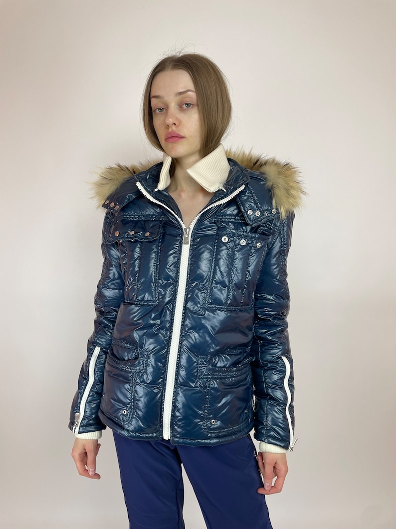 Puede incluir: Una chaqueta acolchada azul con una cremallera blanca y ribetes blancos. La chaqueta tiene una capucha de piel sint&eacute;tica y un bolsillo grande en la parte delantera.