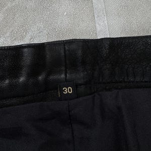 May include: A black leather garment featuring a textured waistband and a small black fabric tag centred on the interior with the number 30 printed in a light gold colour. The interior lining is made of a smooth, solid black fabric material.