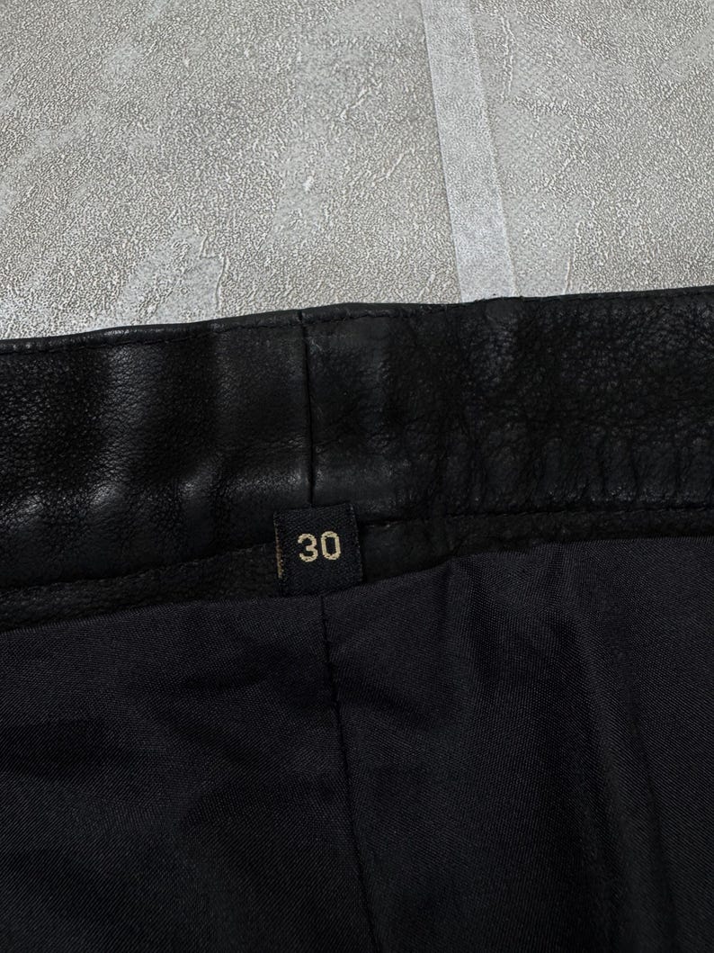 May include: A black leather garment featuring a textured waistband and a small black fabric tag centred on the interior with the number 30 printed in a light gold colour. The interior lining is made of a smooth, solid black fabric material.