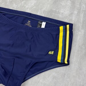May include: Navy blue swim briefs with a yellow and black stripe detail on the side. The briefs feature the H&M logo and the Olympic Collection logo. A classic swimwear item.