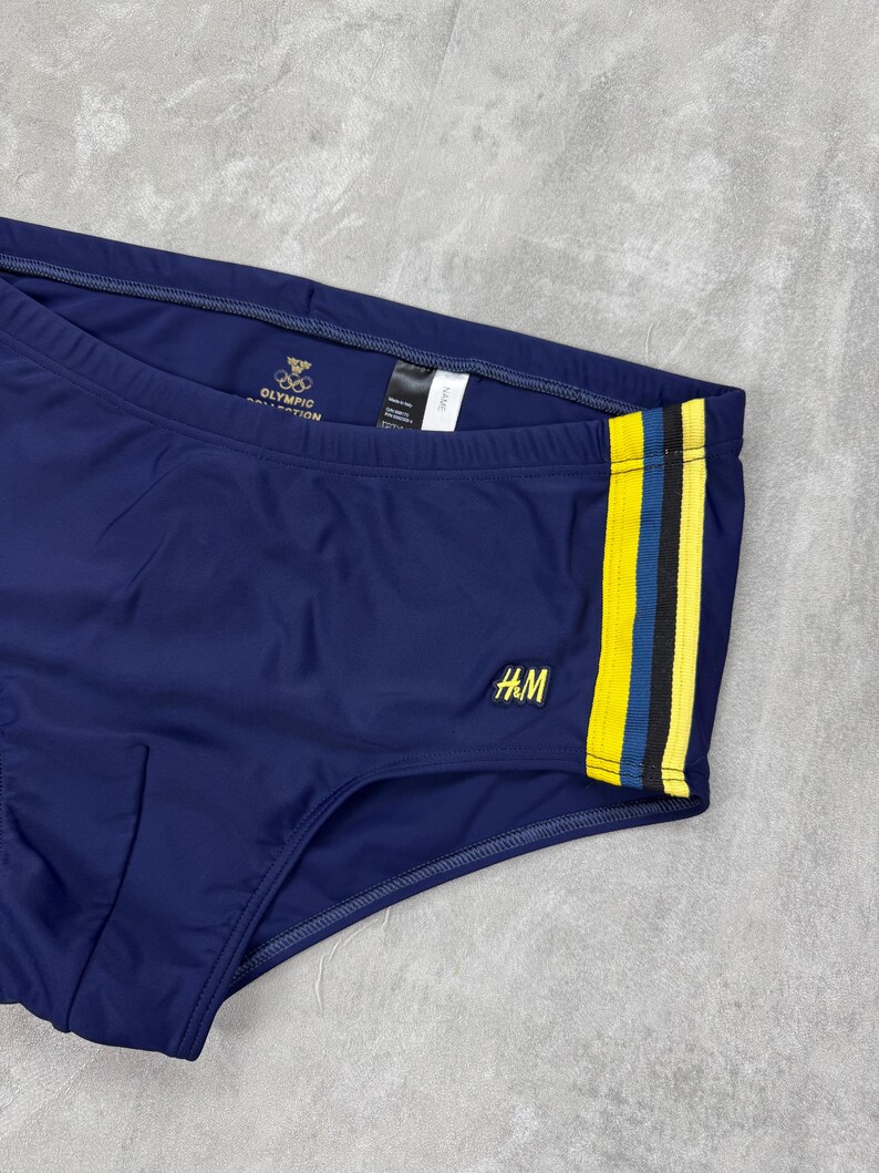 May include: Navy blue swim briefs with a yellow and black stripe detail on the side. The briefs feature the H&M logo and the Olympic Collection logo. A classic swimwear item.