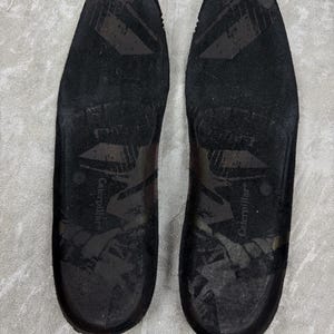 May include: A pair of black insoles with the brand name "Caterpillar" printed on each. The insoles have a textured surface and are designed to fit inside shoes. The image is taken from a top-down perspective.