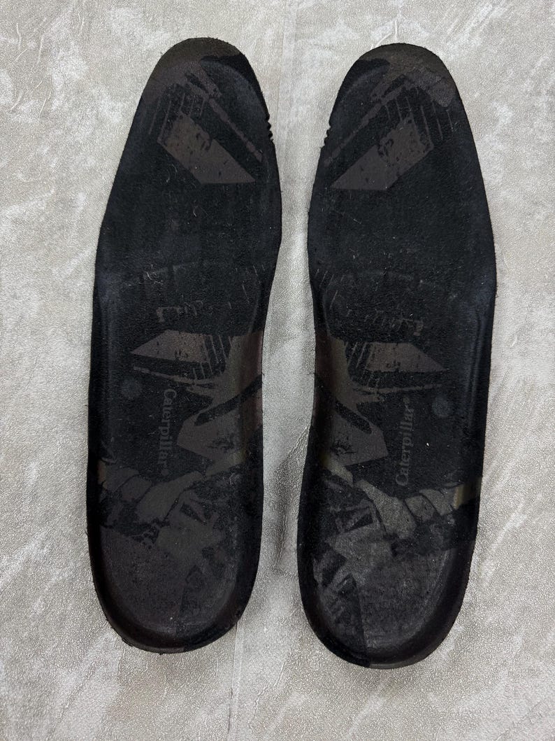May include: A pair of black insoles with the brand name "Caterpillar" printed on each. The insoles have a textured surface and are designed to fit inside shoes. The image is taken from a top-down perspective.