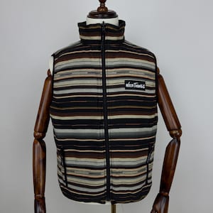 May include: A striped vest with a high collar and a full-length zipper. The vest features horizontal stripes in shades of brown, black, gray, and tan. The "Wild Things" logo is on the chest. The vest has two side pockets.