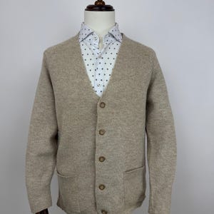 May include: A beige cardigan sweater with a V-neck and button closure. The sweater has two front pockets and is worn over a white button-down shirt with a small, dark pattern. The sweater is displayed on a mannequin.