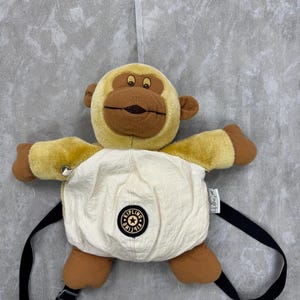 Peut inclure: Un sac à dos en forme de singe en peluche, de couleur beige et marron. Le sac à dos a un corps crème avec un logo circulaire et des bretelles noires. Le visage et les membres du singe sont détaillés avec des accents bruns.