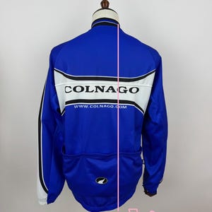 May include: A blue cycling jersey with the brand name "COLNAGO" in white block letters across the back. The jersey has white and black accents, a small logo, and the website address www.COLNAGO.com.