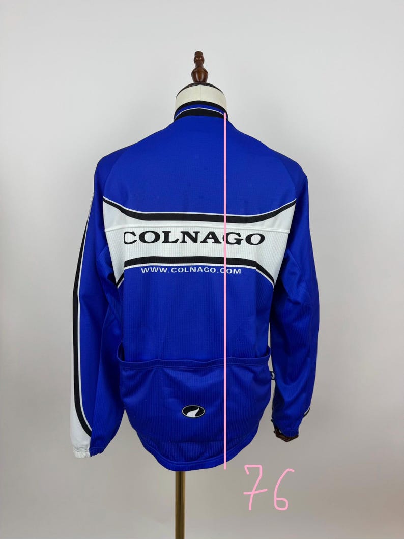 May include: A blue cycling jersey with the brand name "COLNAGO" in white block letters across the back. The jersey has white and black accents, a small logo, and the website address www.COLNAGO.com.
