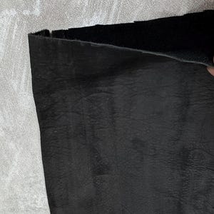 May include: A piece of black leather with a visible grain texture. The material is folded over at the top edge, revealing a smooth, dark underside. The surface displays natural creases and variations in the leather texture across the material.