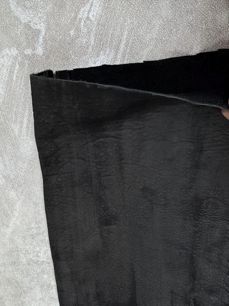 May include: A piece of black leather with a visible grain texture. The material is folded over at the top edge, revealing a smooth, dark underside. The surface displays natural creases and variations in the leather texture across the material.