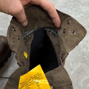 May include: Close-up of a brown leather boot interior. The boot has a suede-like texture and metal eyelets for laces. A yellow tag with text is visible, indicating size 13 USA.