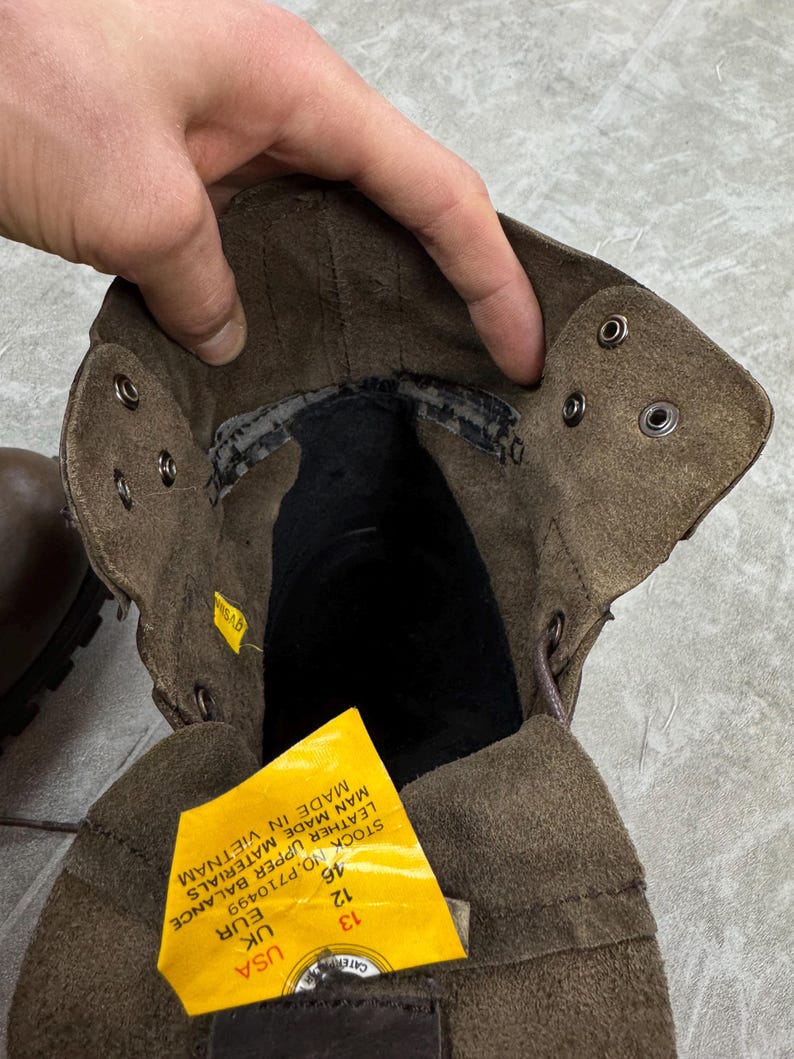 May include: Close-up of a brown leather boot interior. The boot has a suede-like texture and metal eyelets for laces. A yellow tag with text is visible, indicating size 13 USA.
