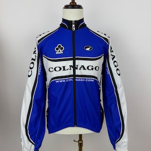May include: A blue, white, and black Colnago cycling jacket with a full-length zipper. The jacket features the Colnago logo and website address. The sleeves are white with black accents. The jacket is displayed on a mannequin.
