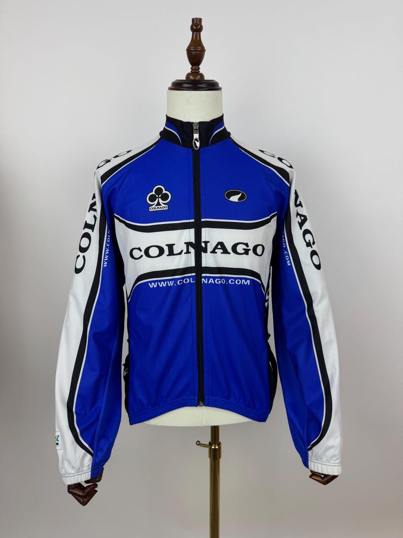 May include: A blue, white, and black Colnago cycling jacket with a full-length zipper. The jacket features the Colnago logo and website address. The sleeves are white with black accents. The jacket is displayed on a mannequin.