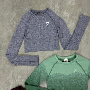 May include: Three cropped long-sleeve athletic tops in shades of blue, green, and burgundy. Each top features a crew neck and a white logo on the chest. The tops are made of a textured, athletic material.