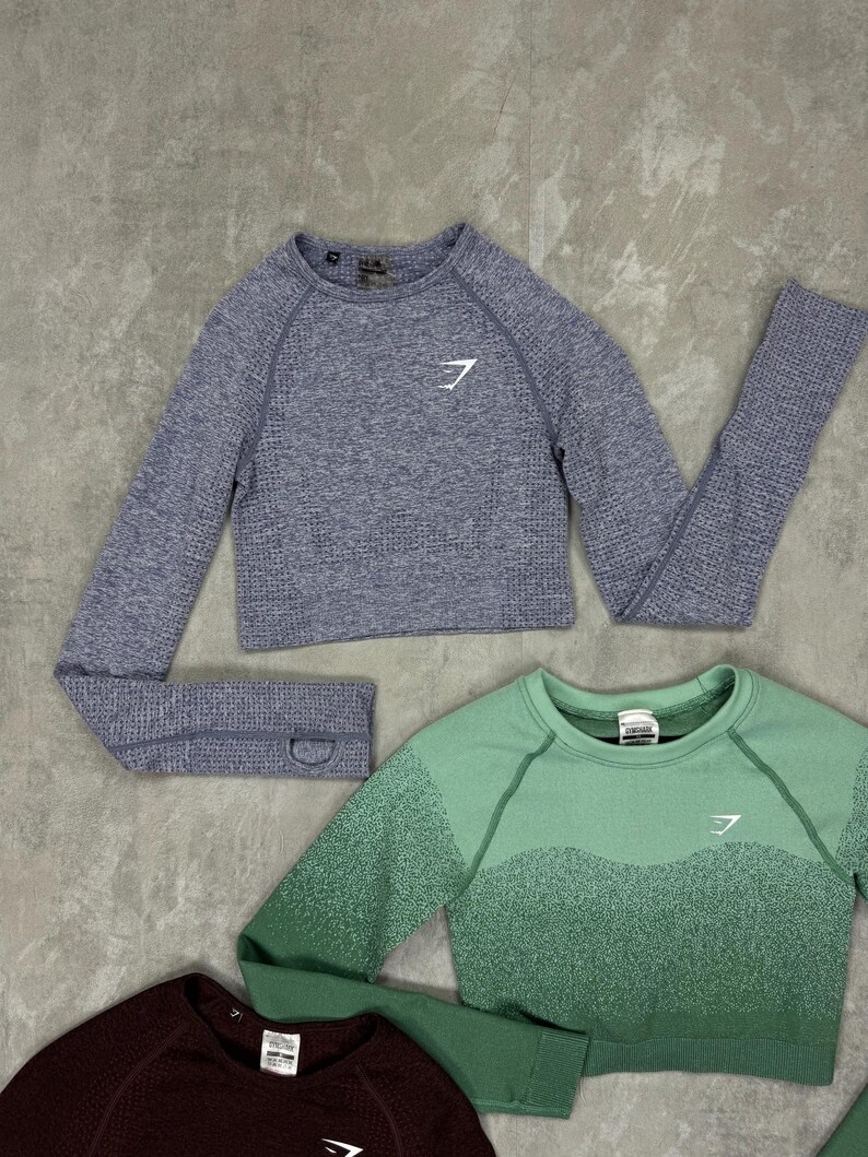 May include: Three cropped long-sleeve athletic tops in shades of blue, green, and burgundy. Each top features a crew neck and a white logo on the chest. The tops are made of a textured, athletic material.