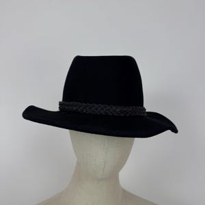 May include: A black fedora-style hat with a wide brim and a braided band. The hat is displayed on a mannequin head against a plain white background. The hat's crown is a medium height.