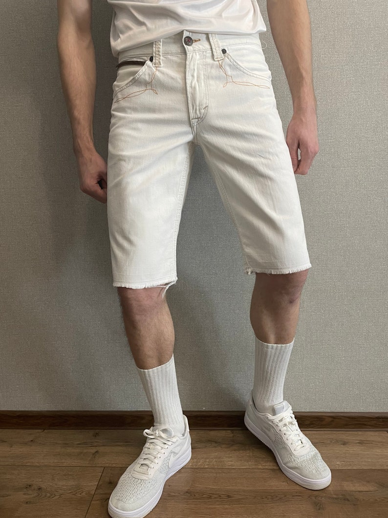 May include: White denim shorts with frayed hems and a brown leather belt. The shorts have a relaxed fit and are worn with white socks and white sneakers.