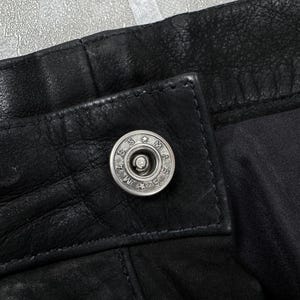 May include: A black leather garment featuring a silver-toned metal snap button closure. The button is circular with the text MASS engraved around the centre, separated by small stars. The leather has a visible grain texture and stitched seams.