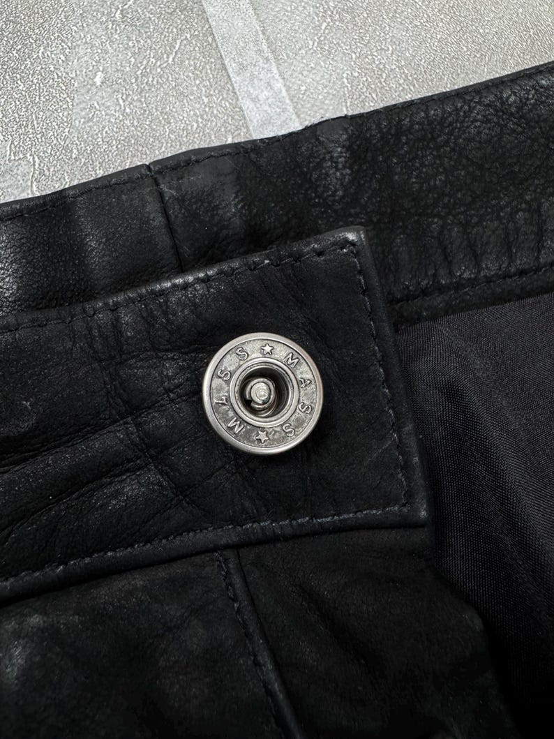 May include: A black leather garment featuring a silver-toned metal snap button closure. The button is circular with the text MASS engraved around the centre, separated by small stars. The leather has a visible grain texture and stitched seams.