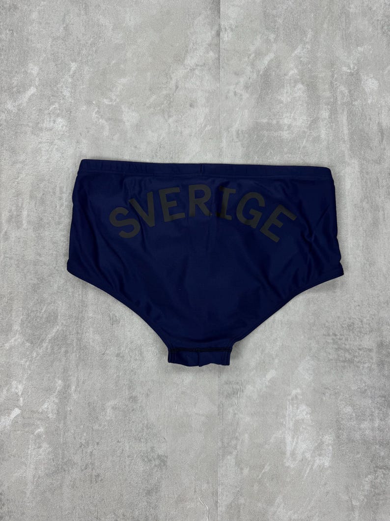 May include: Navy blue swim briefs with the word "SVERIGE" printed across the back in dark green. The swimwear is a solid color and appears to be made of a smooth, possibly synthetic material. The briefs have a classic, brief cut.