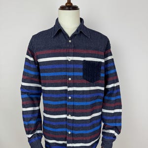 Puede incluir: Camisa de manga larga con botones y rayas horizontales en azul, blanco y burdeos. La camisa tiene cuello azul oscuro y un bolsillo azul oscuro en el pecho. Se muestra en un maniquí.