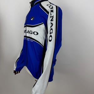 May include: Blue and white cycling jacket with the word "COLNAGO" printed on the sleeve. The jacket has a full-length zipper and a black collar. The jacket has a white panel on the front and sleeves, with black trim. The jacket is displayed on a mannequin.