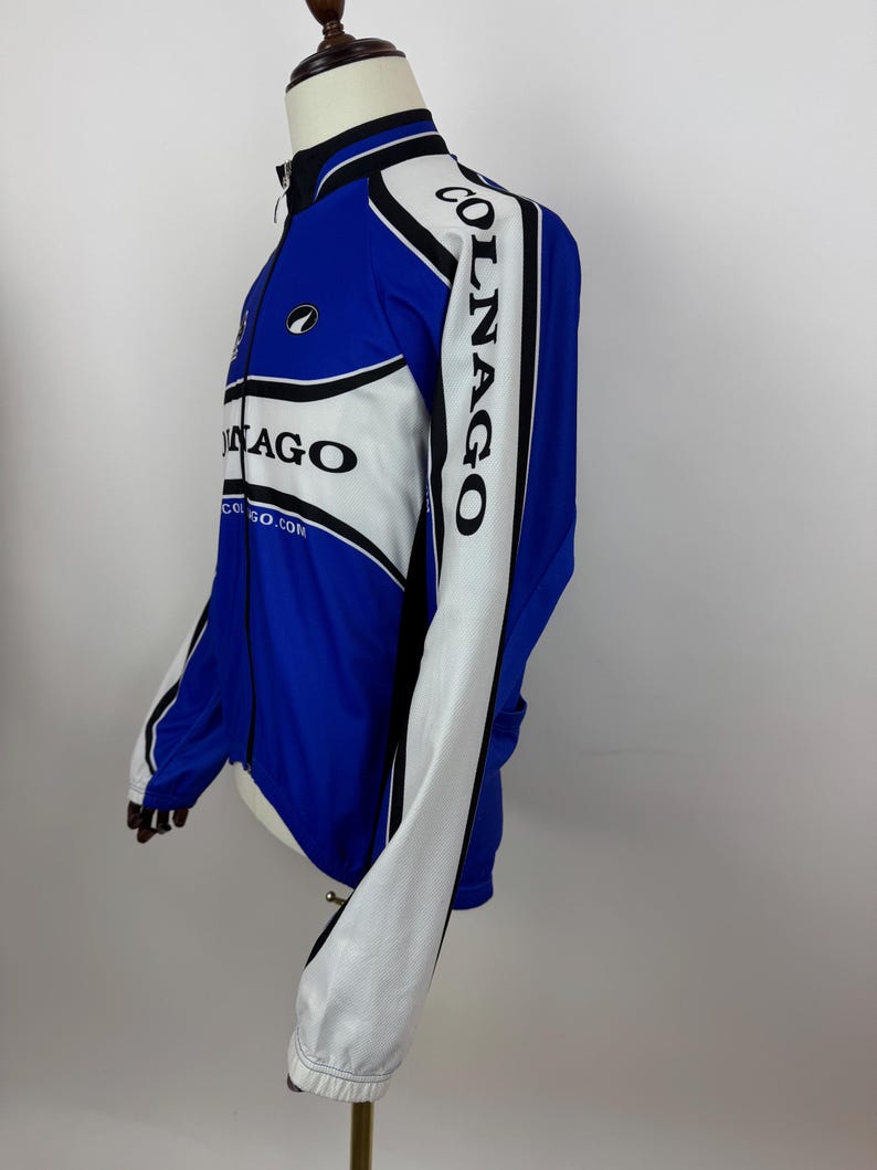 May include: Blue and white cycling jacket with the word "COLNAGO" printed on the sleeve. The jacket has a full-length zipper and a black collar. The jacket has a white panel on the front and sleeves, with black trim. The jacket is displayed on a mannequin.