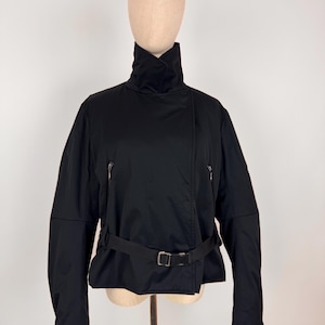 Może przedstawiać: Czarna kurtka z wysokim kołnierzem i paskiem. Kurtka ma ukośne zapięcie na zamek błyskawiczny i dwie małe kieszenie zapinane na zamek. Pasek ma srebrną klamrę. Kurtka jest prezentowana na manekinie.