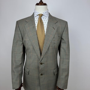 May include: A tailored blazer in a brown, black, and orange check pattern, displayed on a mannequin. The jacket features a notched lapel, two buttons, and two front pockets. A white shirt with a small blue pattern and a gold tie complete the look.