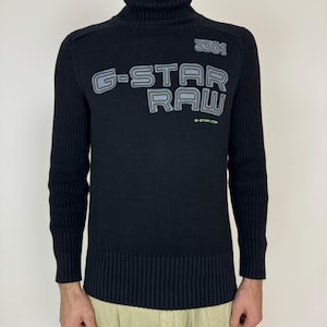 May include: Black turtleneck sweater with a ribbed texture and a G-Star RAW logo in gray and white lettering. The logo is on the front of the sweater and includes the number 3301. The sweater has long sleeves and a ribbed hem.