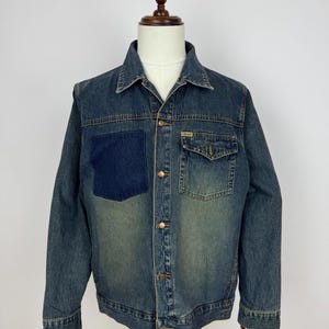 May include: A denim jacket with a dark blue patch on the left chest. The jacket has two front pockets, gold-colored buttons, and a classic collar. The denim is a faded blue with a slightly worn appearance.
