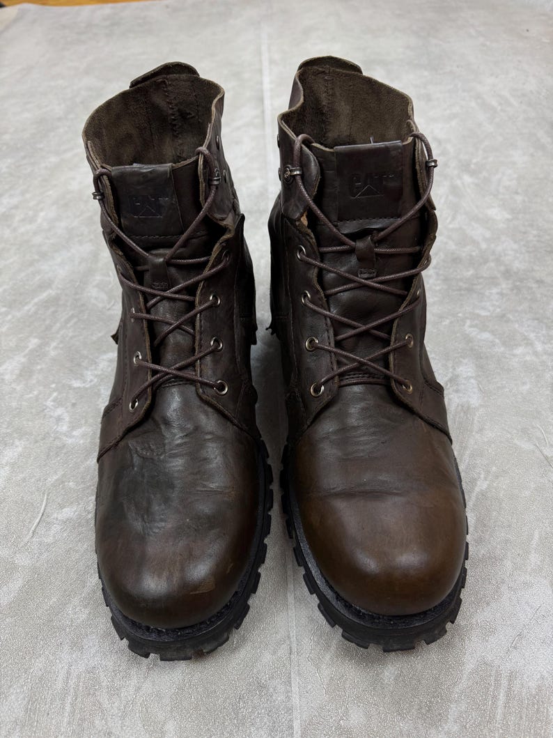 May include: A pair of brown leather work boots with a lace-up design. The boots have a rugged, worn appearance, with visible stitching and a thick, textured sole. The CAT logo is visible on the tongue of the boots.