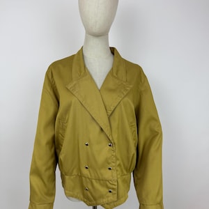 Könnte beinhalten: Senffarbene, zweireihige Jacke mit Reverskragen und silberfarbenen Knöpfen. Die Jacke hat lange Ärmel und eine leicht verkürzte Passform. Das Kleidungsstück wird auf einer Schaufensterpuppe vor einem schlichten weißen Hintergrund präsentiert.