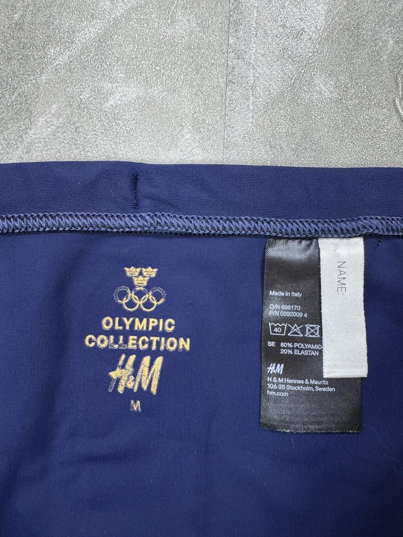 May include: Navy blue athletic wear with gold text reading "OLYMPIC COLLECTION" and the H&M logo. A tag indicates the item is made in Italy and lists the materials as 80% Polyamid and 20% Elastan. The size is medium.