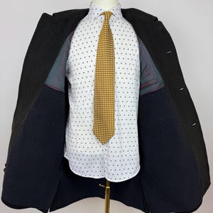 May include: A dark charcoal overcoat is open, revealing a white button-down shirt with a small, repeating pattern. A gold-colored tie with a dotted design is centered. The coat is displayed on a mannequin.