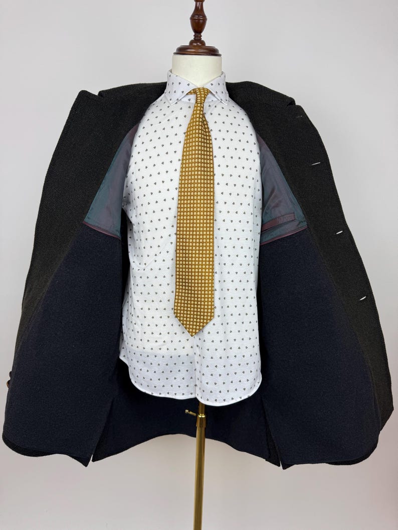 May include: A dark charcoal overcoat is open, revealing a white button-down shirt with a small, repeating pattern. A gold-colored tie with a dotted design is centered. The coat is displayed on a mannequin.