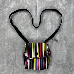 Op de afbeelding: Een kleine, kleurrijke crossbodytas met verticale strepen in rood, geel, wit, zwart en blauw. De tas heeft een zwarte band en een klepsluiting met een zwarte knoop. De achtergrond is een grijze, getextureerde ondergrond.