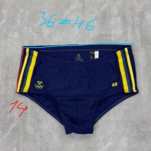 May include: Navy blue swim trunks with yellow and red stripes on the sides. The trunks feature the Olympic rings logo and the H&M logo. The waist measures approximately 36 inches.