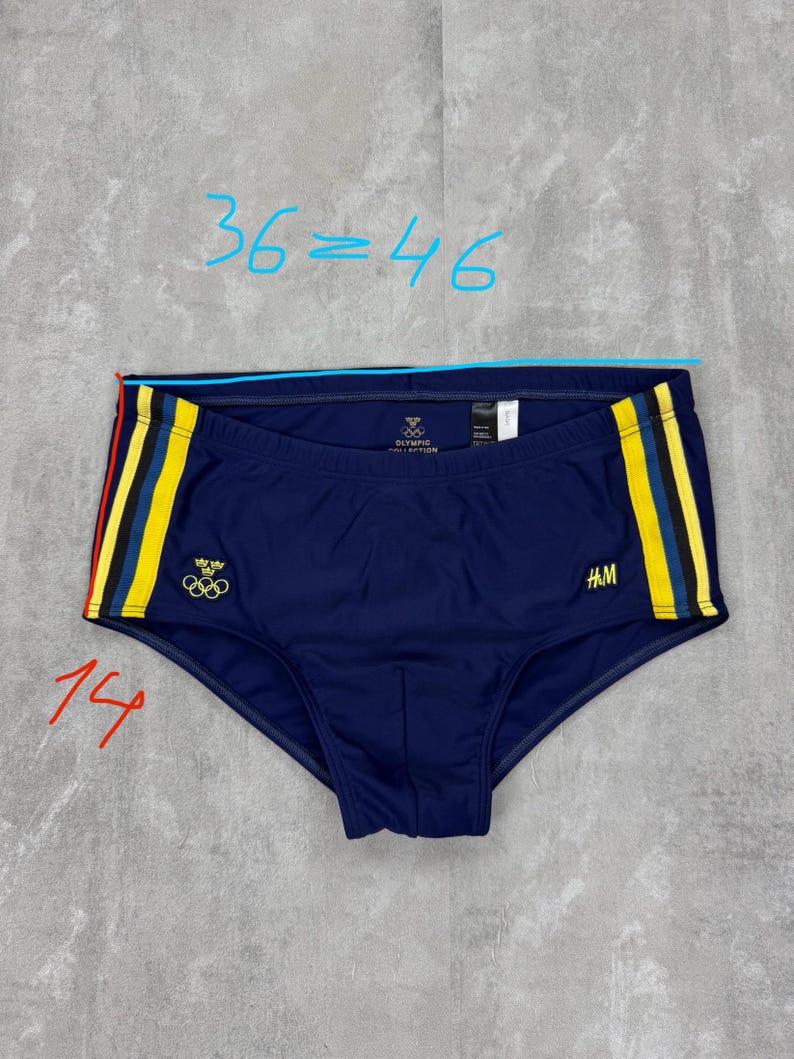 May include: Navy blue swim trunks with yellow and red stripes on the sides. The trunks feature the Olympic rings logo and the H&M logo. The waist measures approximately 36 inches.
