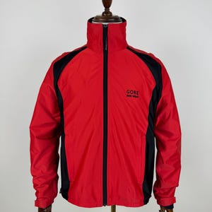 Pode incluir: Casaco Gore Bike Wear vermelho e preto com zíper completo e gola alta. O casaco tem painéis pretos nos ombros e mangas. O logotipo Gore Bike Wear é impresso no peito.